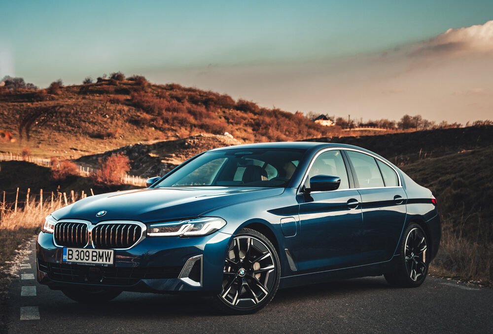 A dark blue BMW sedan is parked on a road in front of rolling hills, with the sky displaying soft clouds and evening light.